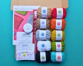 May include: A craft kit in an open cardboard box, containing balls of yarn in various colours, knitting needles, a tape measure, and printed instructions. The yarn colours include brown, red, orange, grey, yellow, white, green, black, and teal. The text "ARTHUR'S Craft Corner" is visible.