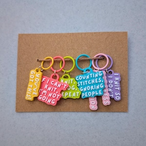 May include: A collection of brightly coloured knitting stitch markers displayed on a brown card. Each marker features a ring in a different colour and a charm with a funny knitting quote, including "You've Got This!" and "I Knit So I Don't Stress."