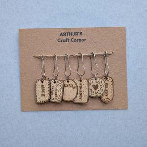May include: Six wooden stitch markers with silver hooks, each shaped like a biscuit and engraved with a word: "Nice", "Custard Cream", "Festive", "Bourbon", a heart, and "Rich Tea". They are attached to a brown card with the text "ARTHUR'S Craft Corner".