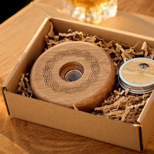 Op de afbeelding: Een houten onderzetter met een Keltisch knoopontwerp en een klein blikje houtpoets. De onderzetter zit in een bruine kartonnen doos met gescheurd papier.