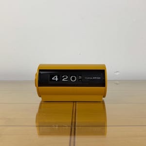 Könnte beinhalten: Eine gelbe Klappuhr mit der Anzeige "4:20". Die Uhr hat eine rechteckige Form mit einem schwarzen Anzeigefenster. Die Uhr steht auf einer Holzoberfläche.