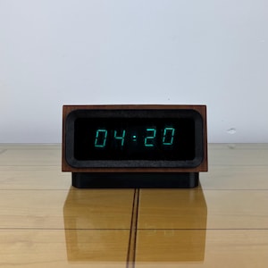 Może przedstawiać: Cyfrowy zegar wyświetlający godzinę 04:20 w zielonych cyfrach. Zegar ma ciemną drewnianą ramę z czarną obwódką. Spoczywa na czarnej podstawie, umieszczonej na jasnej drewnianej powierzchni.