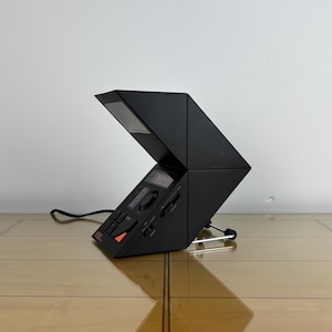 Könnte beinhalten: Ein schwarzes, geometrisch geformtes Vintage-Radiowecker mit einem einzigartigen, eckigen Design. Die Uhr verfügt über eine Digitalanzeige, Bedienelemente und eine kleine Antenne. Das Netzkabel ist sichtbar. Die Uhr steht auf einer Holzoberfläche.