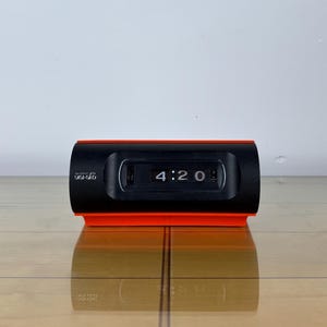 Può includere: Orologio a scatto nero e arancione che mostra le 4:20. L'orologio ha una forma rettangolare con un quadrante nero e una base arancione. Il marchio "DIGI-GLO" è stampato sul lato sinistro del quadrante. L'orologio è su una superficie di legno.