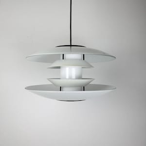 Könnte beinhalten: Eine weiße, mehrstufige Pendelleuchte mit schwarzem Kabel und Akzenten. Die Leuchte hat eine Reihe von sich überlappenden, tellerförmigen Schirmen, die ein geschichtetes Design erzeugen. Die Leuchte hängt vor einem schlichten weißen Hintergrund.