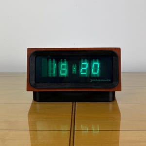 Pode incluir: Um relógio digital vintage exibindo 18:20 em algarismos verdes brilhantes. O relógio tem uma moldura retangular de madeira com uma face e base pretas. O texto "Электроника" é visível no visor.