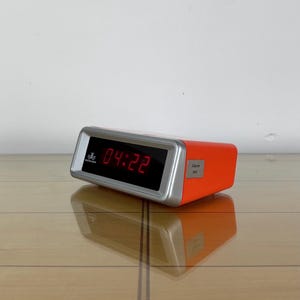 Peut inclure: Réveil numérique orange avec un cadre argenté, affichant l'heure 04:22 en rouge. Le réveil est posé sur une surface en bois. Le nom de la marque est visible sur le cadran. Dimensions: environ 15 cm de long.