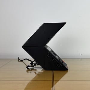 May include: A black, angular stereo system with a hinged top, resting on a chrome stand. The device has a control panel with buttons and a display. A power cord extends from the back.