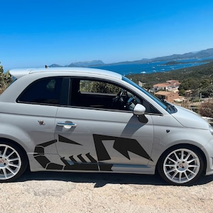 Può includere: Un'auto compatta grigio chiaro con una grafica di scorpione nero sul lato. L'auto ha ruote bianche e uno spoiler posteriore. Lo sfondo presenta un cielo blu e un paesaggio costiero.