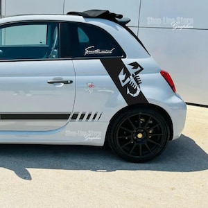 Può includere: Una Fiat 500 argentata con strisce da corsa nere e una grafica a scorpione sul lato. L'auto ha ruote nere e un tetto nero.