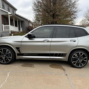 Op de afbeelding: Zilveren SUV met zwarte accenten, waaronder zwarte velgen en getinte ramen. Het voertuig heeft een zwarte streep langs de zijkant met de tekst "X3 Performance". De auto staat geparkeerd op een betonnen oprit voor een huis.