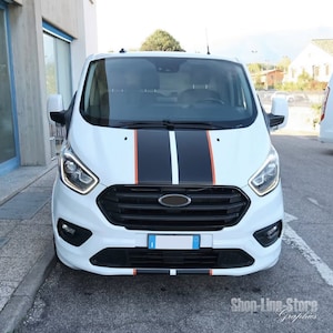 Peut inclure: Fourgon Ford Transit blanc avec des bandes de course noires et orange sur le capot. Le fourgon a une calandre noire et un pare-chocs noir. Le fourgon est garé devant un bâtiment.