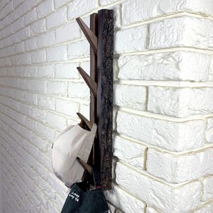 May include: A dark brown wooden coat rack with four hooks, mounted on a white brick wall. A white hat and a dark blue jacket are hanging on the hooks.