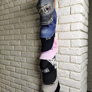 May include: A wall mounted hat rack with seven baseball caps hanging on it. The caps are in various colors and styles, including black, blue, pink, and gray. The wall is white brick.