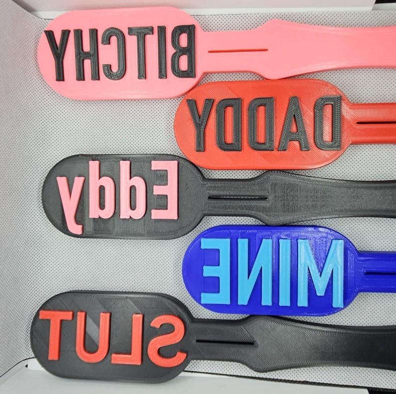 May include: A collection of paddle-shaped objects in various colors, each with a different word in bold, raised letters. The colors include pink, red, black, blue, and the words are "BITCH", "DADDY", "EGGY", "MINE", and "SLUT".