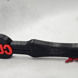 May include: A black paddle-shaped object with the word "CUSTOM" in red letters. The paddle has a handle and a hole at the end with a red cord attached. The object appears to be 3D-printed.