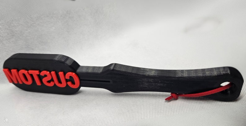 May include: A black paddle-shaped object with the word "CUSTOM" in red letters. The paddle has a handle and a hole at the end with a red cord attached. The object appears to be 3D-printed.
