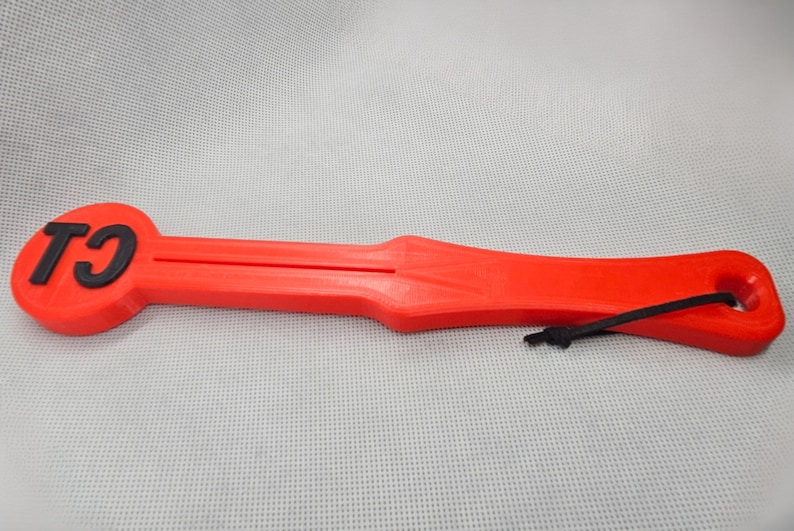 May include: A bright red, elongated tool with a circular end featuring the black letters "TJ". The tool has a handle and a hole at the opposite end, with a black cord attached. The item appears to be 3D-printed.