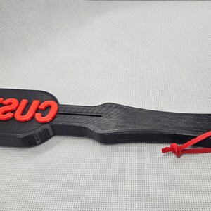 May include: A black paddle-shaped object with the red word "CUSTOM" on the top. The paddle has a handle with a hole and a red string attached. The object is on a light gray surface.