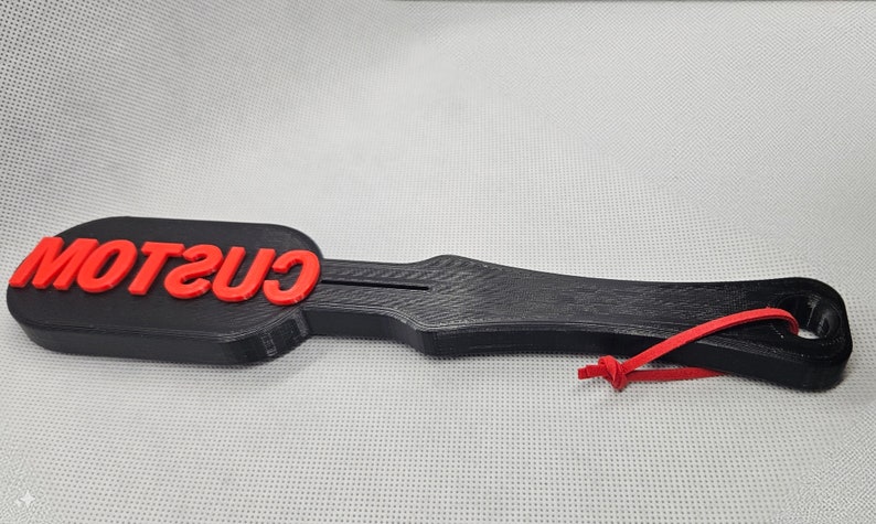 May include: A black paddle-shaped object with the red word "CUSTOM" on the top. The paddle has a handle with a hole and a red string attached. The object is on a light gray surface.