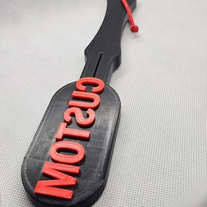 May include: A black paddle with the word "CUSTOM" in red letters. The paddle has a handle with a red cord attached. The paddle is made of a 3D-printed material.