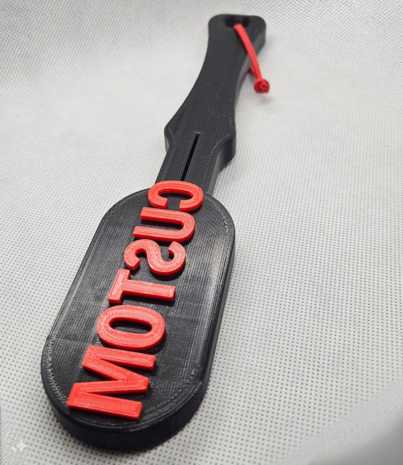 May include: A black paddle with the word "CUSTOM" in red letters. The paddle has a handle with a red cord attached. The paddle is made of a 3D-printed material.