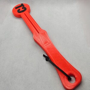 May include: A bright red, 3D-printed tool with the text "G1" in black on a circular end. The tool has a long, slender handle with a central groove and a black cord attached. The overall design is geometric and functional.