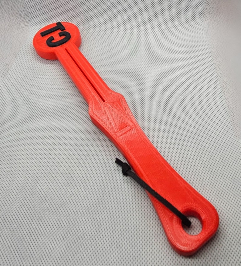 May include: A bright red, 3D-printed tool with the text "G1" in black on a circular end. The tool has a long, slender handle with a central groove and a black cord attached. The overall design is geometric and functional.