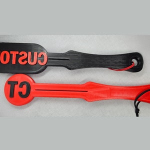 May include: Two paddle-shaped objects, one black and one red, with red text. The black paddle has the word "CUSTOM" in red. The red paddle has the letters "CT" in red. Both have a hole at the end with a cord.