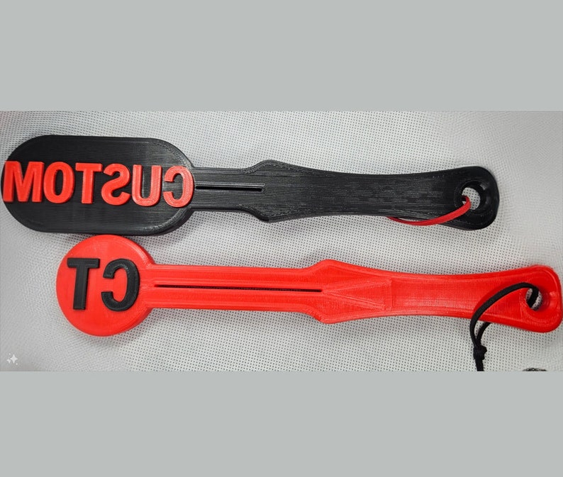 May include: Two paddle-shaped objects, one black and one red, with red text. The black paddle has the word "CUSTOM" in red. The red paddle has the letters "CT" in red. Both have a hole at the end with a cord.