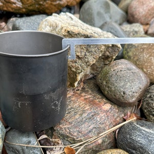 Pode incluir: Uma caneca de acampamento de metal cinza escuro com uma alça prateada. A caneca é cilíndrica com uma pequena alça na lateral. A alça é presa à caneca com um gancho. A caneca está sobre um leito de pedras.