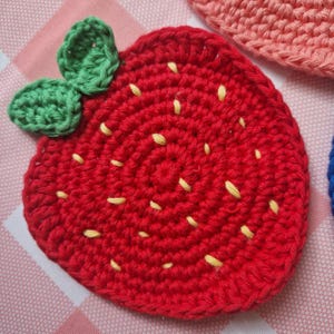 May include: A handmade, red crocheted strawberry coaster with green leaves and yellow seed details. The coaster is round and has a textured surface, perfect for protecting tables from hot or cold drinks. The strawberry design adds a touch of whimsy.