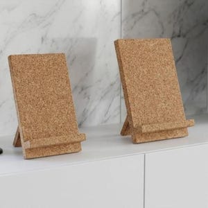 May include: Two black candlestick holders and two cork phone stands are displayed on a white surface. The stands are rectangular with a small ledge to hold a phone. A flat-screen TV is in the background.