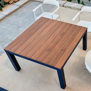 Op de afbeelding: Een vierkante houten tafel met een zwart metalen frame en vier witte stoelen. De tafel heeft een houtnerfpatroon en de stoelen hebben een eenvoudig ontwerp.