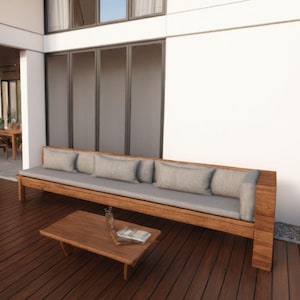 Puede incluir: Un patio exterior de madera con un sofá gris, una mesa de café y un puf gris. El patio está hecho de tablones de madera y tiene una vista de una casa moderna con grandes ventanas.