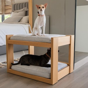 以下が含まれることがあります： 2 段のウッド製のペットベッド。白い小型犬が上の段に座り、黒い猫が下の段に寝ています。ベッドは天然木で作られており、薄いグレーのクッションが付いています。
