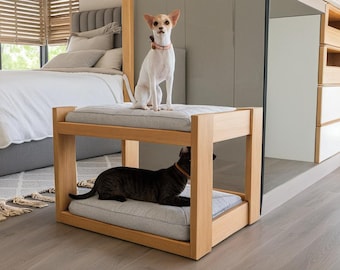 May include: A wooden pet bed with two levels. A small white dog is sitting on the top level and a black cat is lying on the bottom level. The bed is made of natural wood and has a light grey cushion.