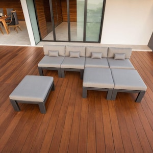 May include: A gray sectional sofa with multiple cushions sits on a wooden deck. The sofa is made up of individual pieces that can be arranged in different configurations. The deck is made of wood planks and is surrounded by a grassy area.