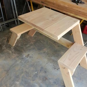 Può includere: Un tavolo pieghevole in legno con due panche. Il tavolo è in legno chiaro e ha un design semplice. Le panche sono anch'esse in legno chiaro e hanno un design semplice.