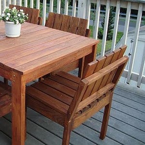 May include: A wooden outdoor dining set with a rectangular table and four chairs. The table and chairs are made of dark brown wood and have a natural finish. The set is on a wooden deck.