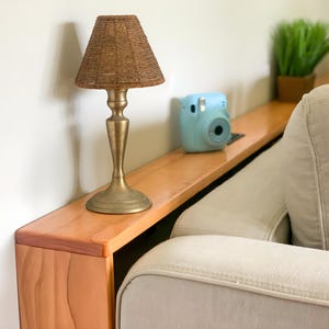 Op de afbeelding: Een kleine, decoratieve lamp met een gouden voet en een geweven lampenkap staat op een houten plank. Een lichtblauwe camera en een kleine potplant staan ook op de plank, die aan een lichtgekleurde bank is bevestigd.