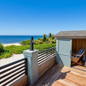 May include: A wooden deck with a view of the sea. A small, light blue shed with a wooden door and a window is on the deck. A white lounge chair is inside the shed. The deck has a black railing and a wooden floor.
