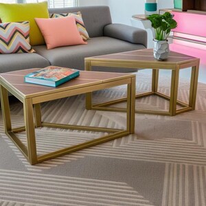 Könnte beinhalten: Zwei goldene dreieckige Couchtische mit einem grauen Sofa und bunten Kissen im Hintergrund. Der Tisch links hat ein Buch darauf. Die Tische stehen auf einem gemusterten Teppich.