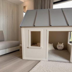 Pode incluir: Uma casinha de brincar para crianças com telhado cinza e paredes brancas. A casa tem uma janela e uma porta aberta. No interior, há um tapete pequeno, uma almofada branca e um bicho de pelúcia redondo e branco. A casinha está numa sala de estar moderna.