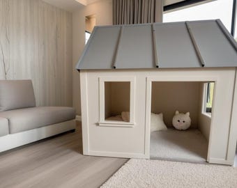 Puede incluir: Una casita de juegos para niños con techo gris y paredes blanquecinas. La casa tiene una ventana y una puerta abierta. En el interior hay una alfombra pequeña, una almohada blanca y un peluche redondo y blanco. La casita está en un salón moderno.