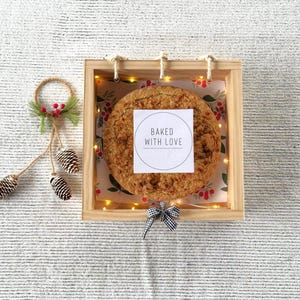 Può includere: Una scatola di legno con una finitura marrone chiaro contiene una torta rotonda ricoperta di briciole con un'etichetta bianca su cui è scritto "Baked with Love". La scatola è decorata con un fiocco bianco e nero e una ghirlanda di luci.