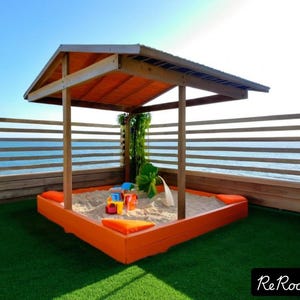 Puede incluir: Un arenero de madera con techo, ubicado en una terraza con vista al mar. El arenero está lleno de arena y tiene algunos juguetes coloridos en su interior. La terraza está hecha de madera y tiene una superficie de césped artificial verde.