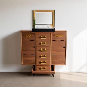 May include: A wooden cabinet with a mirror and gold-colored hardware. The cabinet has multiple drawers and doors, and is set against a white wall. The mirror is rectangular with a gold frame.