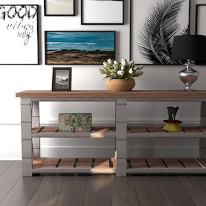 以下が含まれることがあります： 白い木製のコンソールテーブルで、天板は茶色で、棚が3つあります。テーブルの上段には白いシェードの黒いランプ、中段には白い花の入った花瓶、下段には小さな額縁に入った写真があります。テーブルは、白の壁の前にあり、黒と白の額縁に入った写真が飾られています。