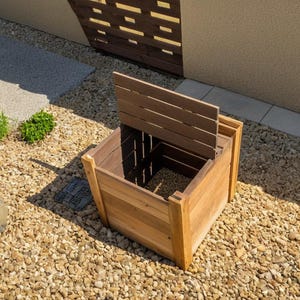Pode incluir: Um contentor de compostagem de madeira com uma tampa articulada que está aberta. O contentor é feito de madeira natural e tem um aspeto rústico. O contentor está situado numa superfície de cascalho.